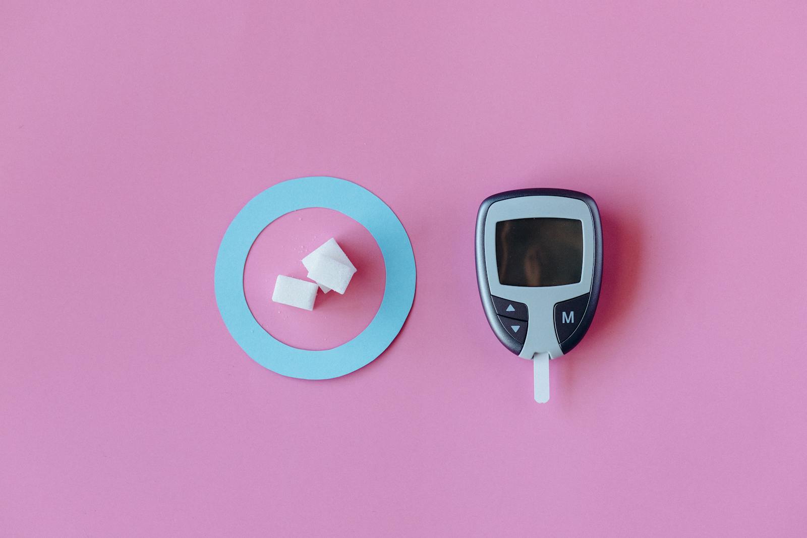 Flat lay of glucometer with sugar cubes on pink background, symbolizing diabetes awareness.